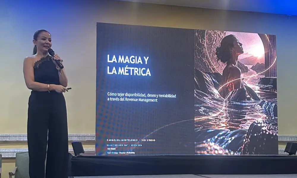 Presenter in a black sleeveless top giving a speech next to a large presentation screen. The screen reads "LA MAGIA Y LA MÉTRICA" with an abstract, light-streaked illustration.