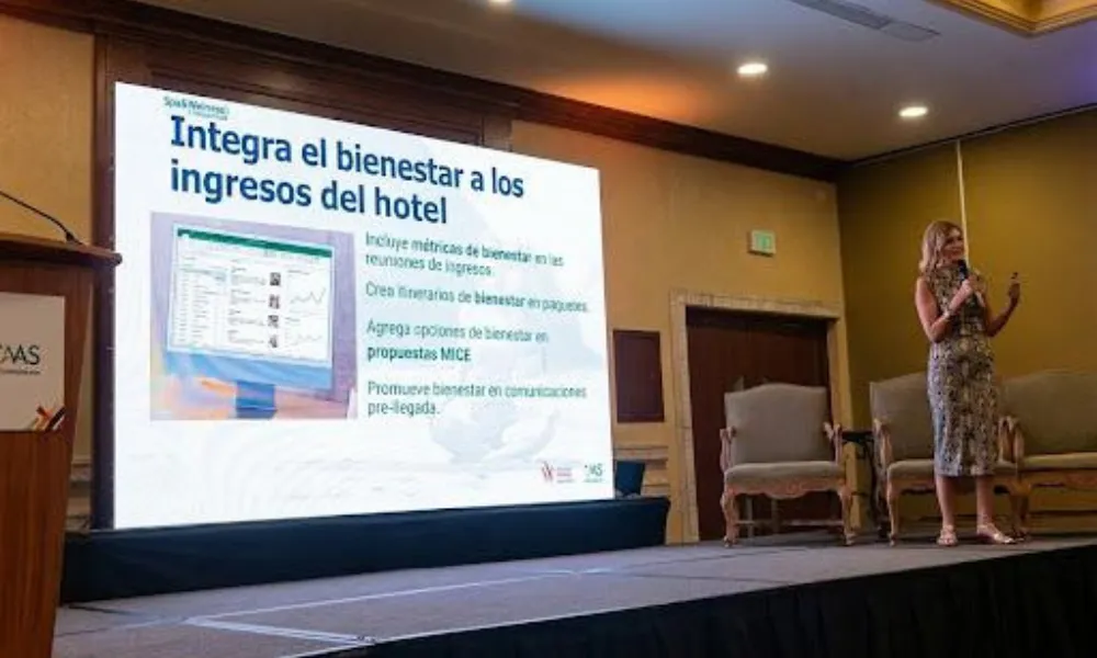 Diapositiva de presentación titulada "Integra el bienestar a los ingresos del hotel." Muestra cuatro estrategias clave para la integración. Una presentadora está de pie a la derecha.